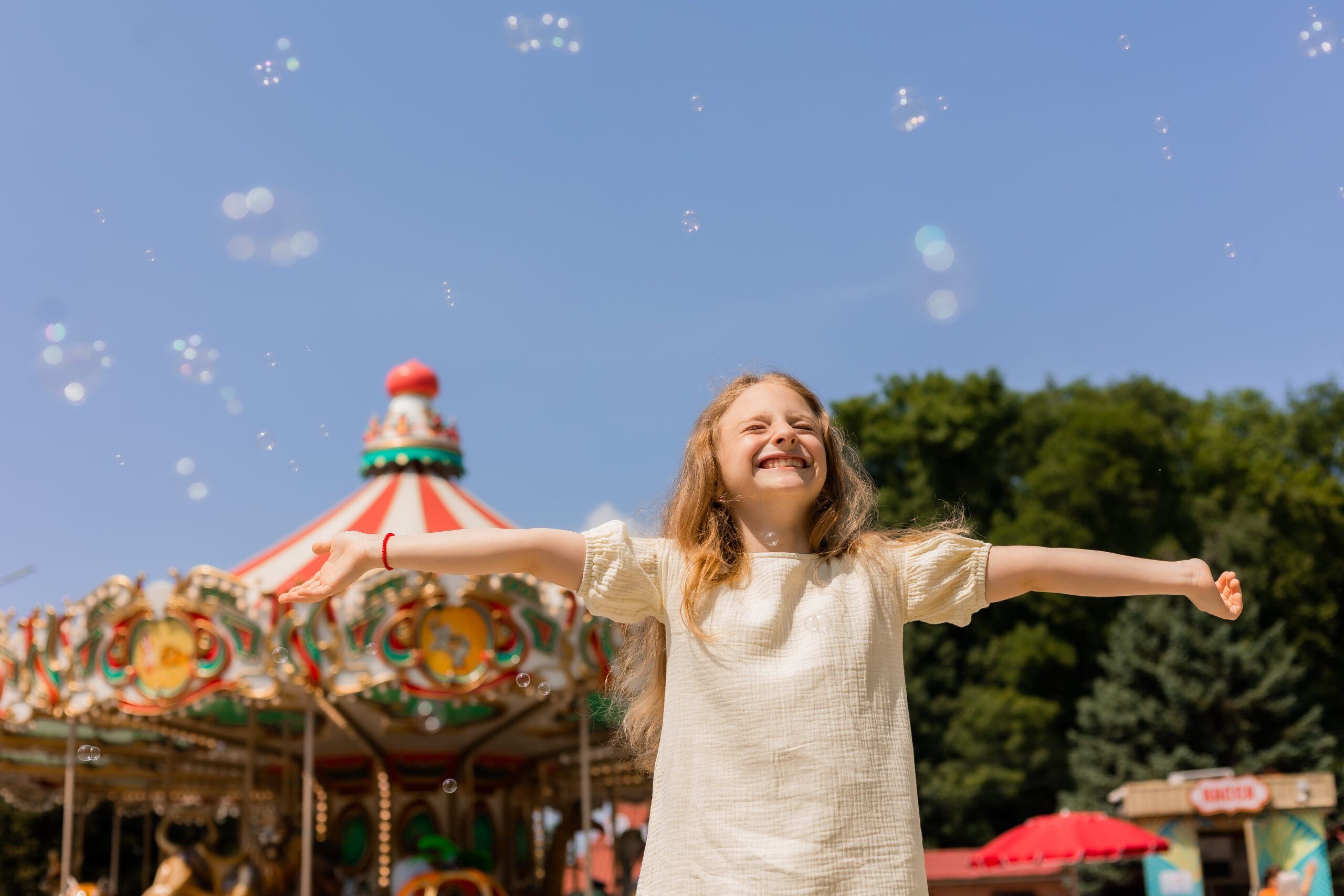 happy little girl is blowing bubbles in front of a carousel at an amusement park